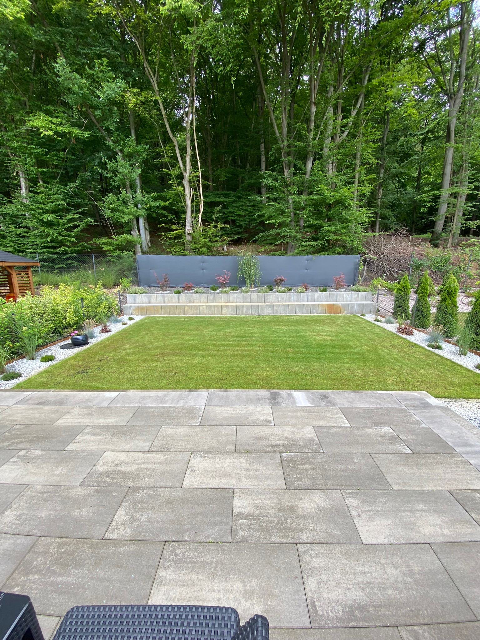 Starannie utrzymany trawnik z równo przystrzyżoną trawą, obrzeża wysypane białym grysem, w tle ozdobne krzewy i drzewa, widoczny fragment tarasu z szarych płyt.