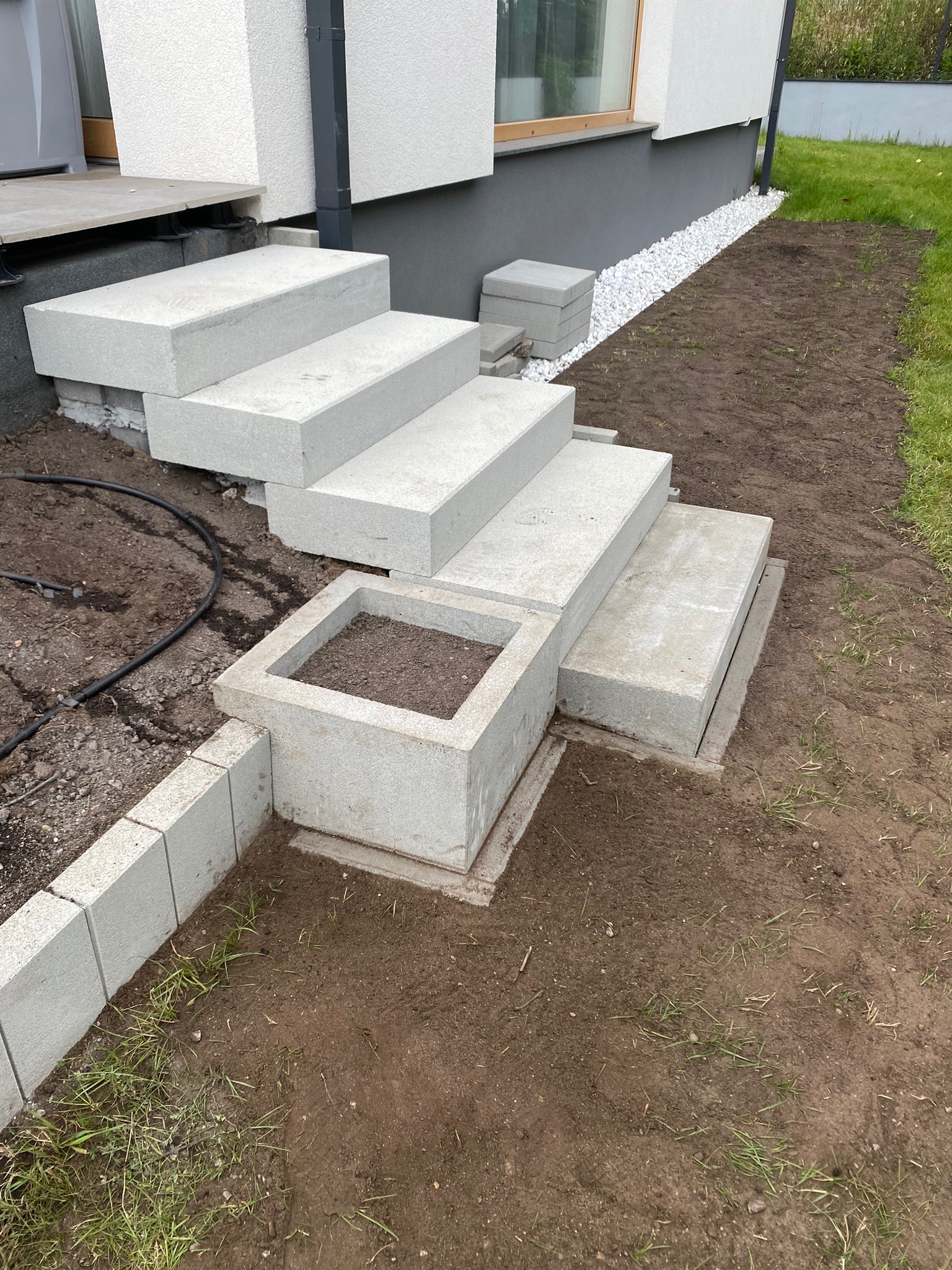 Betonowe schody wejściowe w trakcie budowy, z donicą również z betonu, ziemia przygotowana do nasadzeń, widoczne białe kamienie przy fundamencie domu.