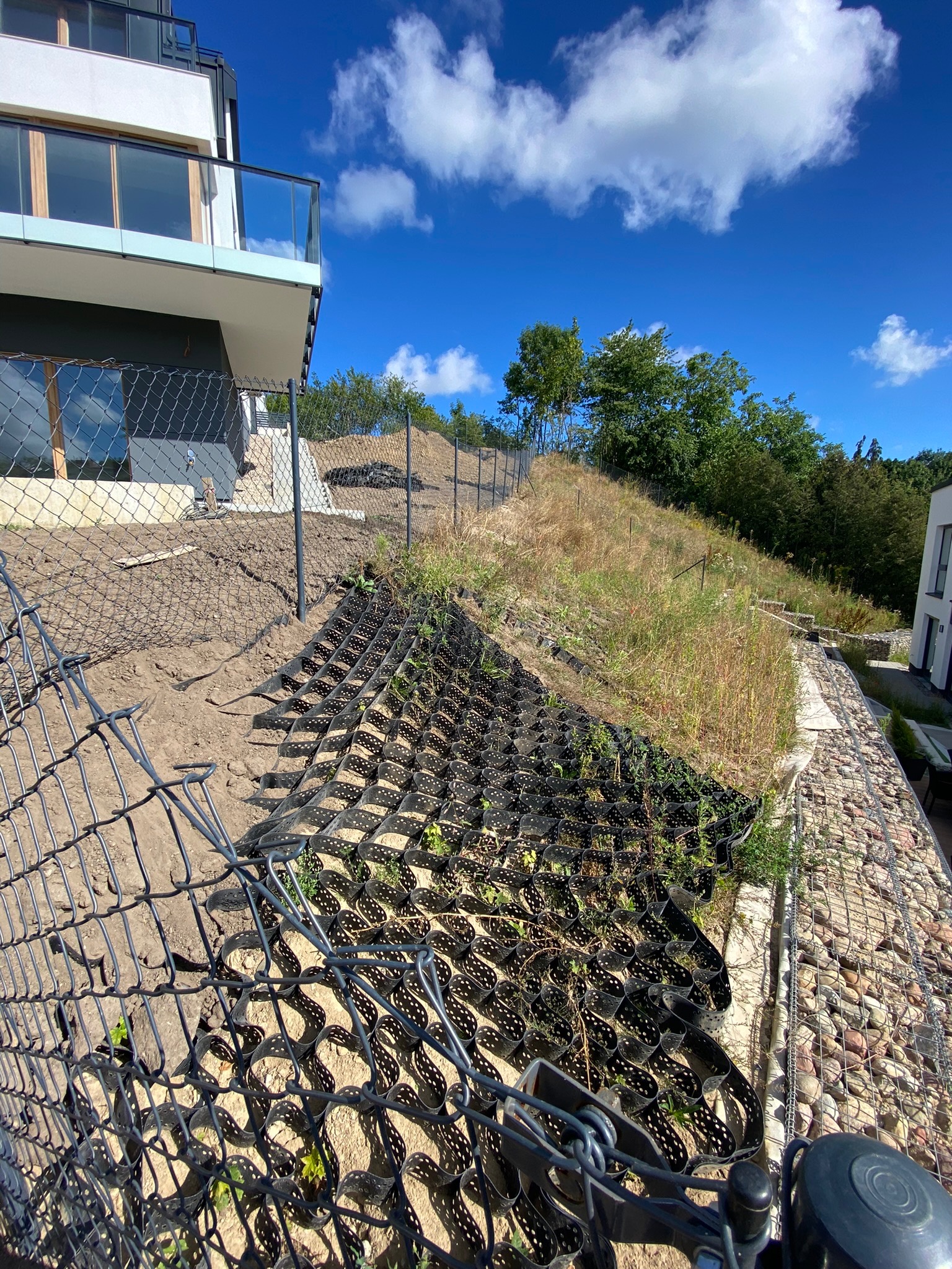 Siatka geokomórkowa na zboczu wzgórza, częściowo obsadzona roślinami, zabezpieczająca przed erozją, widok z dołu na nowoczesny dom i błękitne niebo.