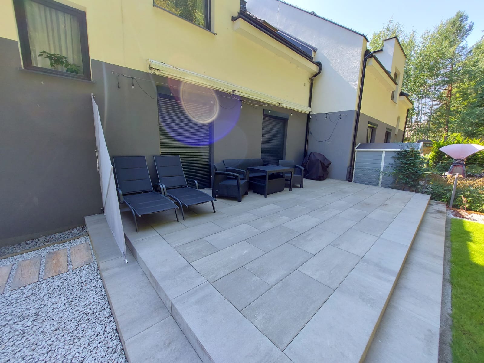 Nowoczesny taras z szarych płyt, wyposażony w czarne leżaki i meble ogrodowe, przylegający do domu z elementami w odcieniach szarości i beżu, otoczony zielenią.