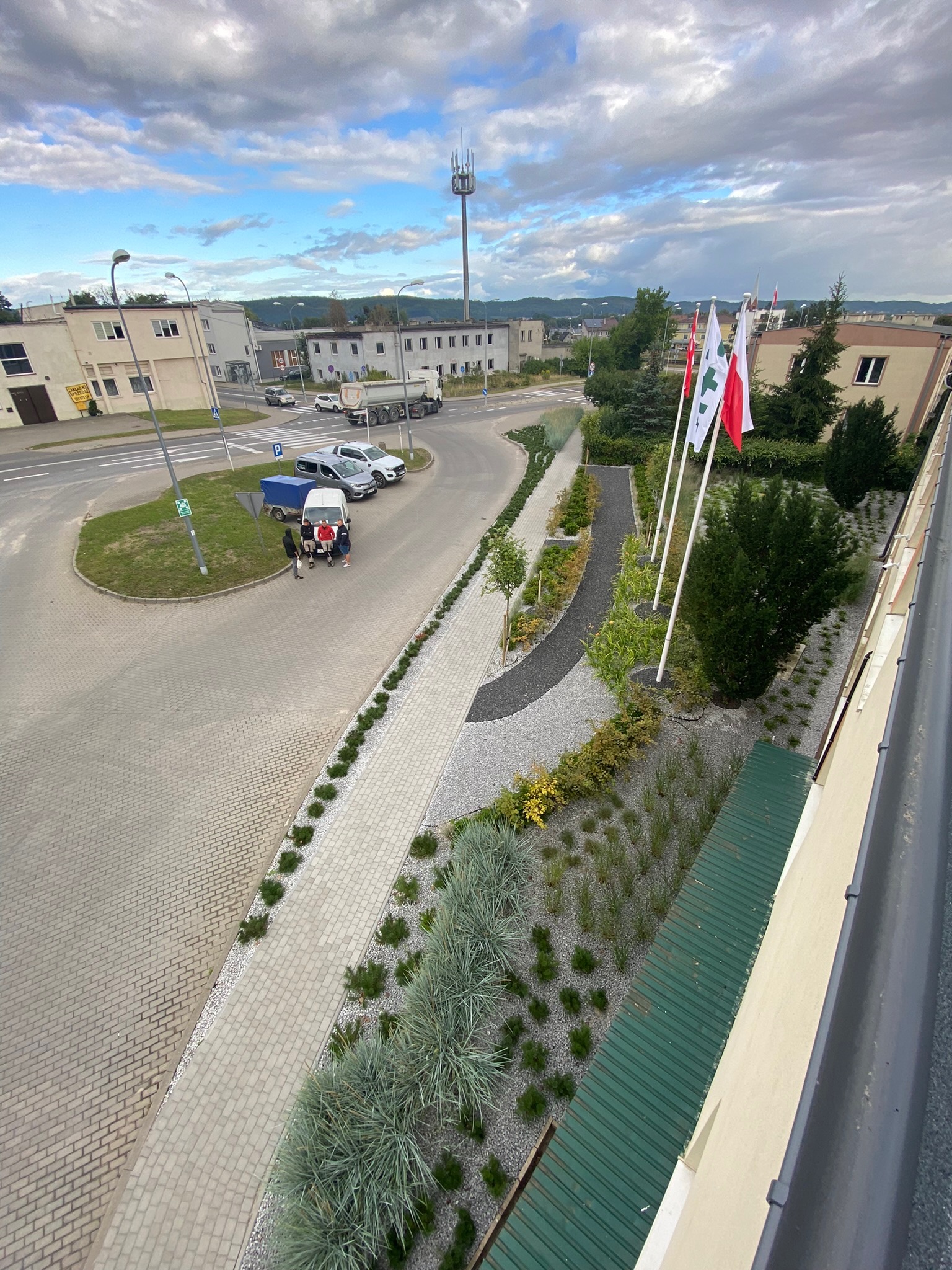 Widok z góry na nowo zaprojektowany teren zielony z chodnikiem, rabatami z roślinami ozdobnymi i flagami, otoczony budynkami i ulicą.