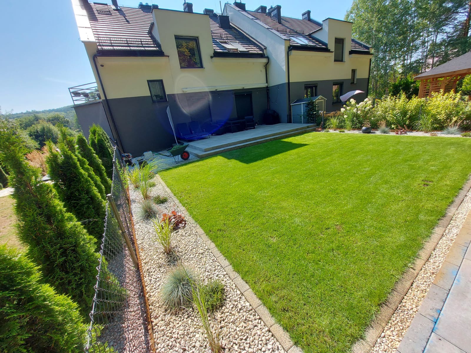 Starannie utrzymany trawnik przylegający do nowoczesnego domu z tarasem, obrzeża z jasnych kamieni i nasadzenia ozdobnych traw, widok z góry.