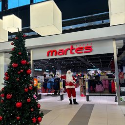 Selfcare Sonia Zgrabczyńska - Święty Mikołaj w czerwonym stroju stoi przed wejściem do sklepu sportowego Martes Sport, obok choinki ozdobionej czerwonymi bombkami, w galerii handlowej.