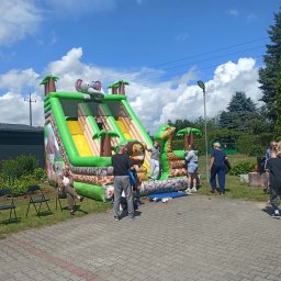 Selfcare Sonia Zgrabczyńska - Duża, zielono-żółta dmuchana zjeżdżalnia z motywami safari (słoń, żyrafa, małpa) w plenerze. Dzieci bawią się na konstrukcji, dorośli obserwują obok, na tle błękitnego nieba z chmurami.