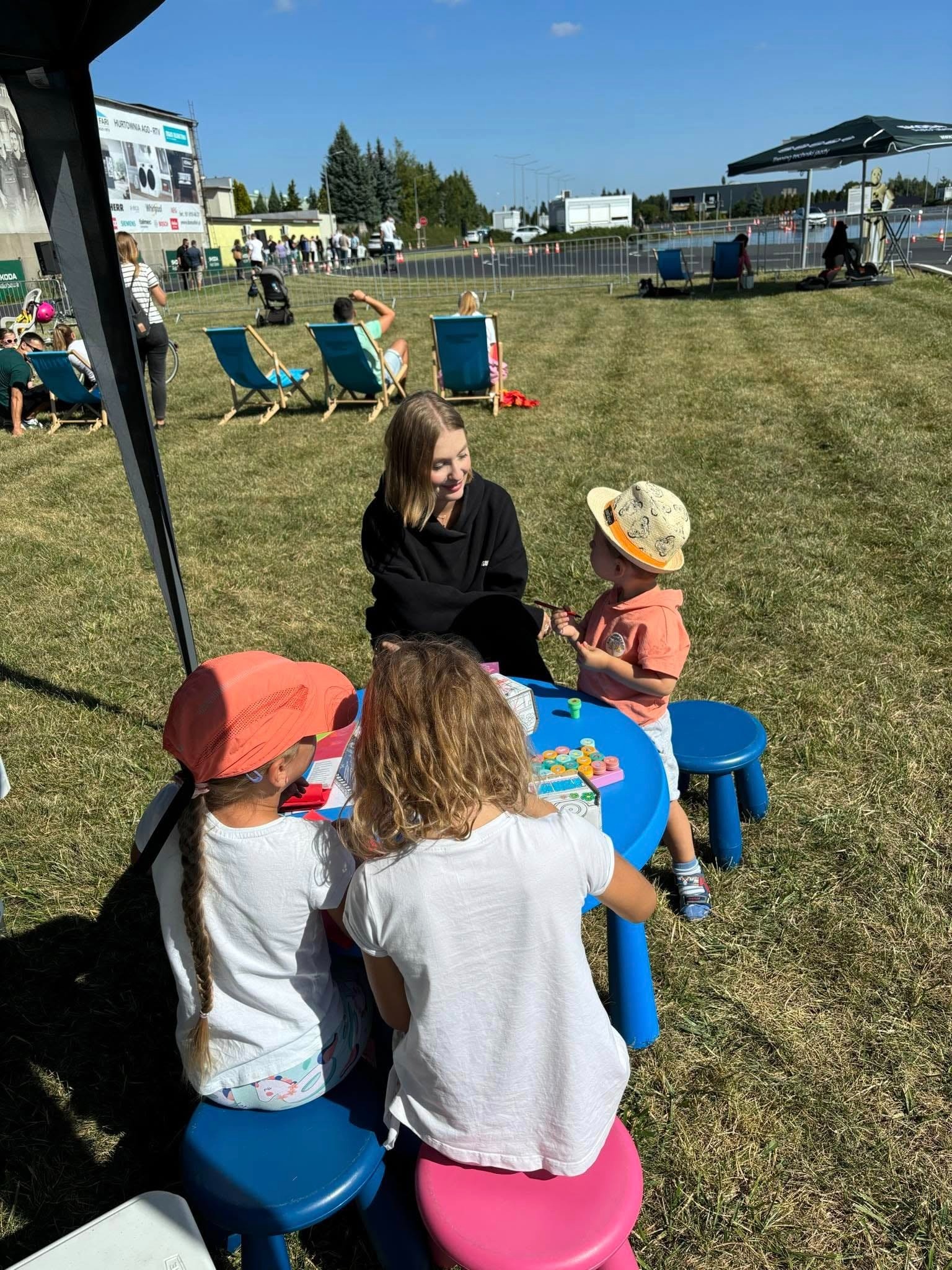 Kobieta w czarnej bluzie uśmiecha się do dzieci bawiących się przy niebieskim stoliku na trawie podczas eventu plenerowego. Dzieci siedzą na kolorowych stołeczkach. W tle widoczni ludzie i reklamy.