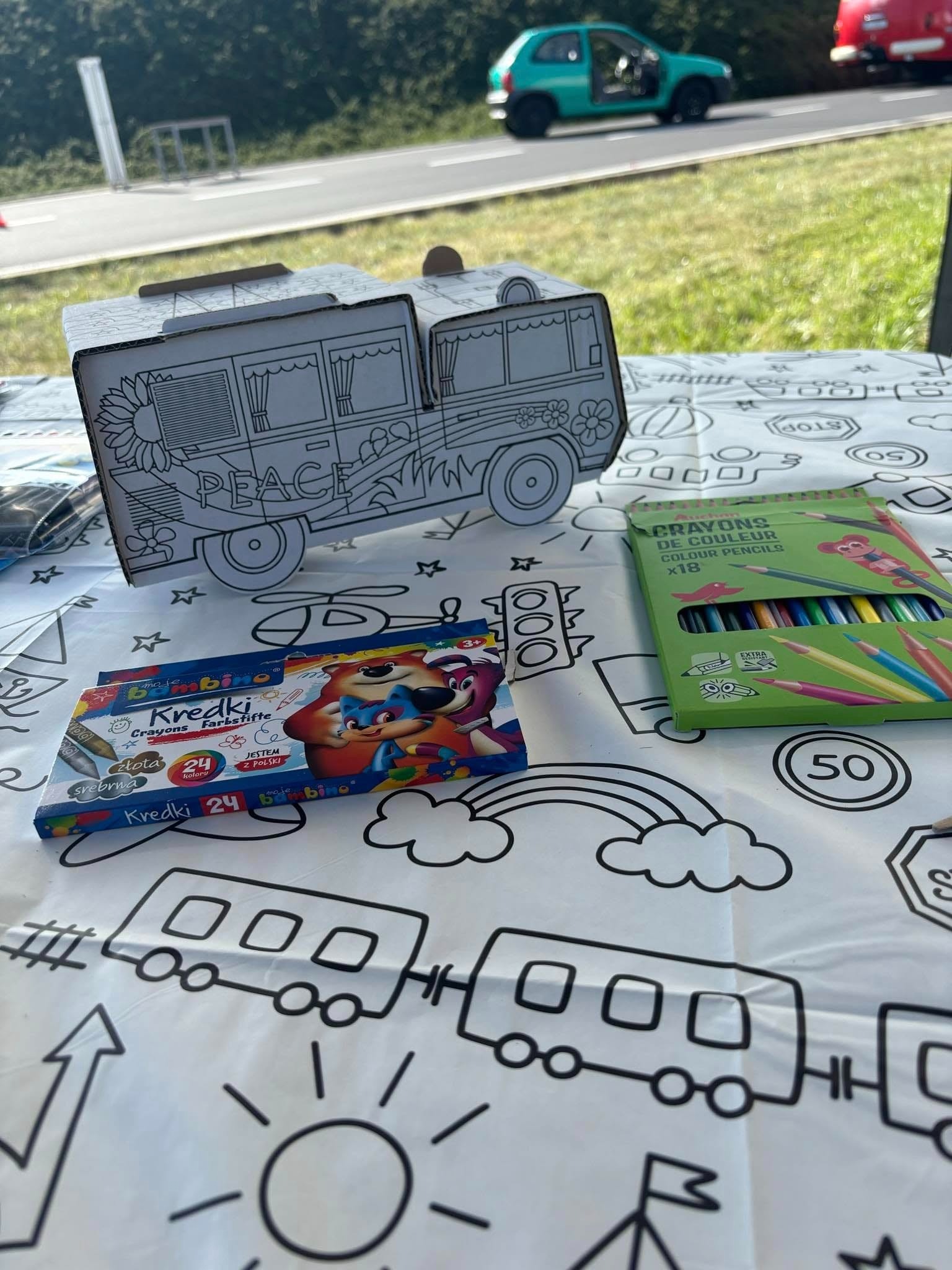 Stół z kolorowanką w formie kampera do samodzielnego pokolorowania, obok pudełka kredek Bambino i Auchan, w tle fragment ulicy z zaparkowanymi samochodami.