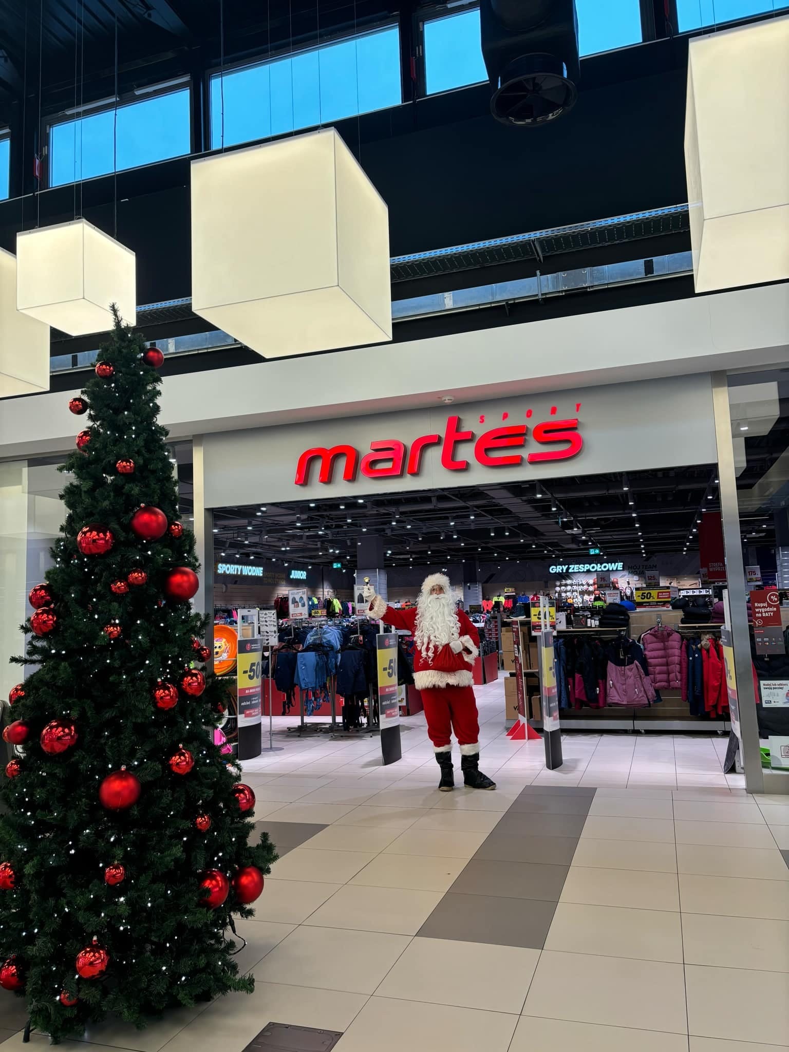 Święty Mikołaj w czerwonym stroju stoi przed wejściem do sklepu sportowego Martes Sport, obok choinki ozdobionej czerwonymi bombkami, w galerii handlowej.