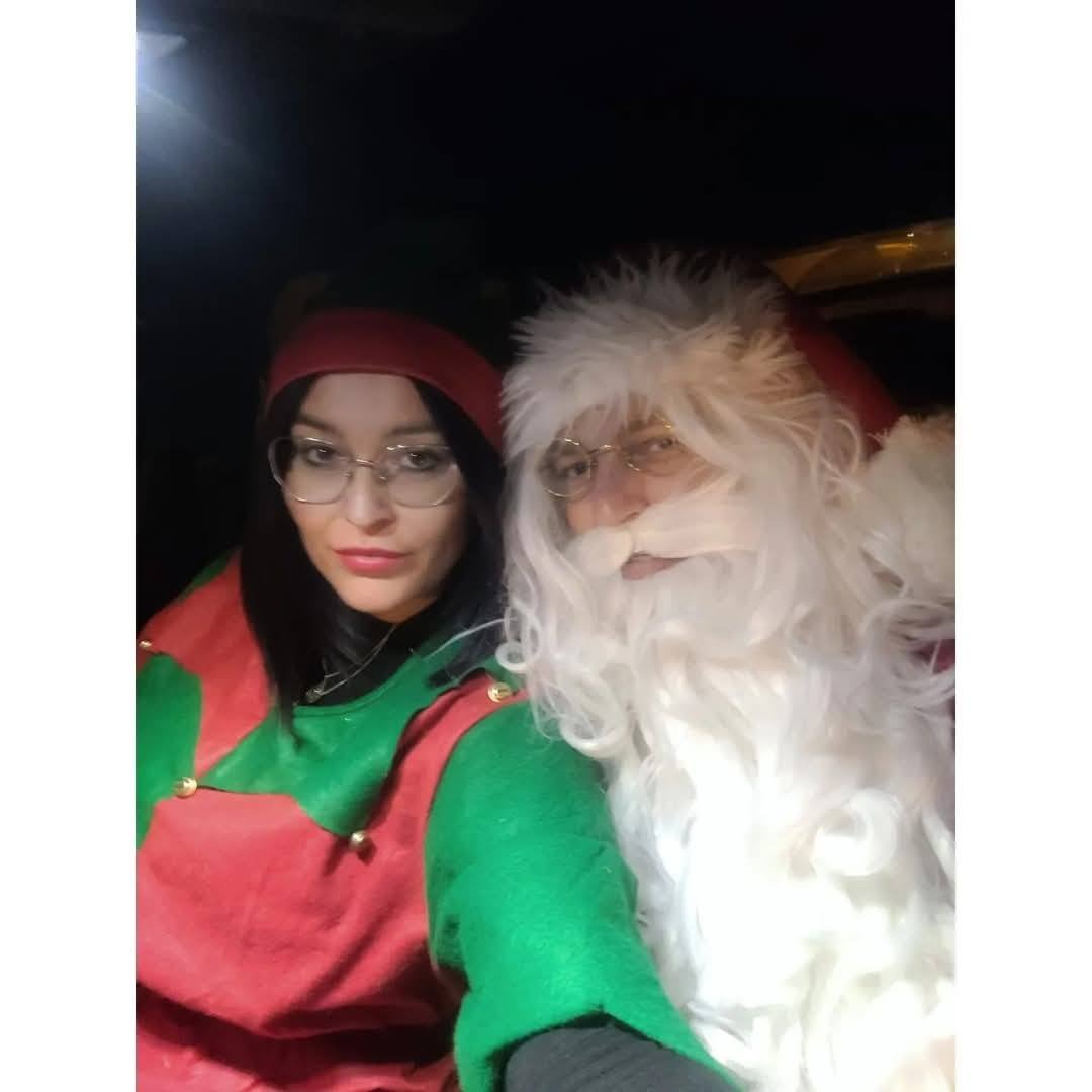 Kobieta w kostiumie elfa i mężczyzna przebrany za Świętego Mikołaja pozują do selfie, oboje uśmiechnięci i w okularach, słabe oświetlenie.