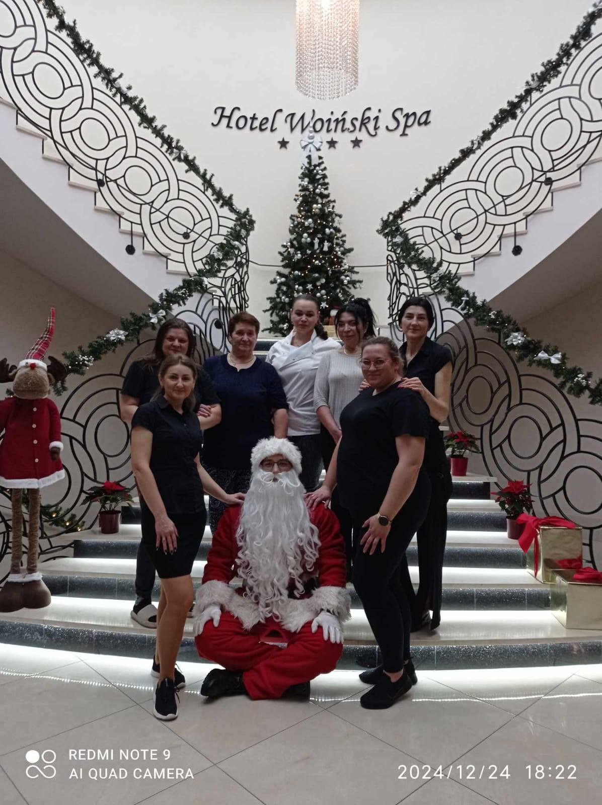 Grupa kobiet i mężczyzna przebrany za Świętego Mikołaja pozują na schodach Hotelu Woinski Spa, ozdobionych świątecznymi dekoracjami, w tle choinka i ozdobny żyrandol.