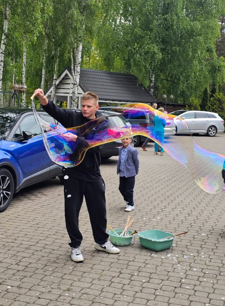 Mężczyzna tworzy gigantyczne bańki mydlane na zewnątrz, obok stoi chłopiec w marynarce. W tle samochody i drzewa. Bańki mienią się kolorami tęczy.