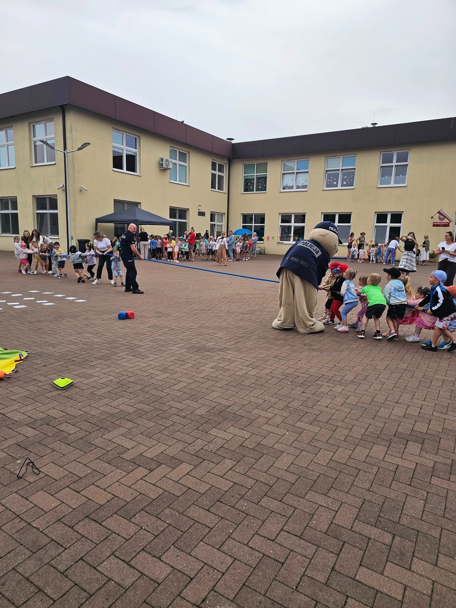 Dzieci i maskotka policjanta biorą udział w przeciąganiu liny na ceglanym placu. W tle budynek szkoły i grupa obserwujących osób.