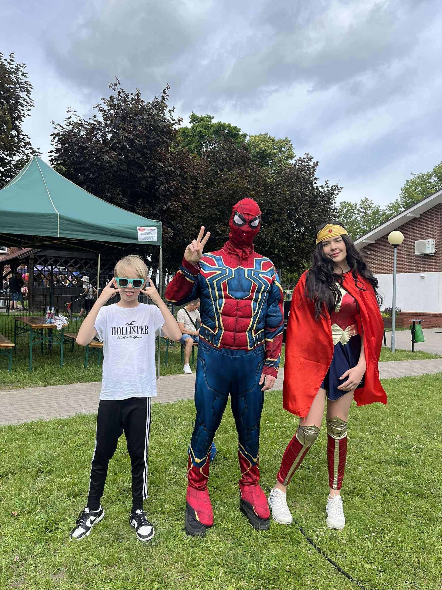 Animatorzy w kostiumach: Spiderman, Wonder Woman i dziecko w okularach, pozują na trawie. W tle zielony namiot i budynek z cegły.