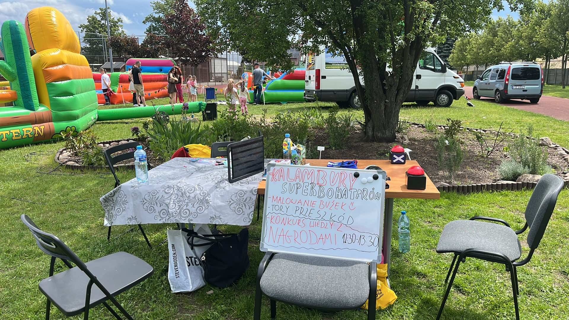 Stanowisko z tablicą informującą o atrakcjach dla dzieci: malowanie buziek, tory przeszkód, konkurs wiedzy z nagrodami. W tle dmuchane zamki i bawiące się dzieci.