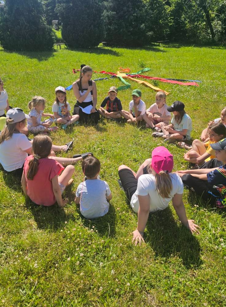 Animator prowadzi zajęcia na trawie dla grupy dzieci siedzących w kręgu. Kolorowe wstążki dekoracyjne. Dzieci w różnym wieku, zrelaksowane, słuchają.