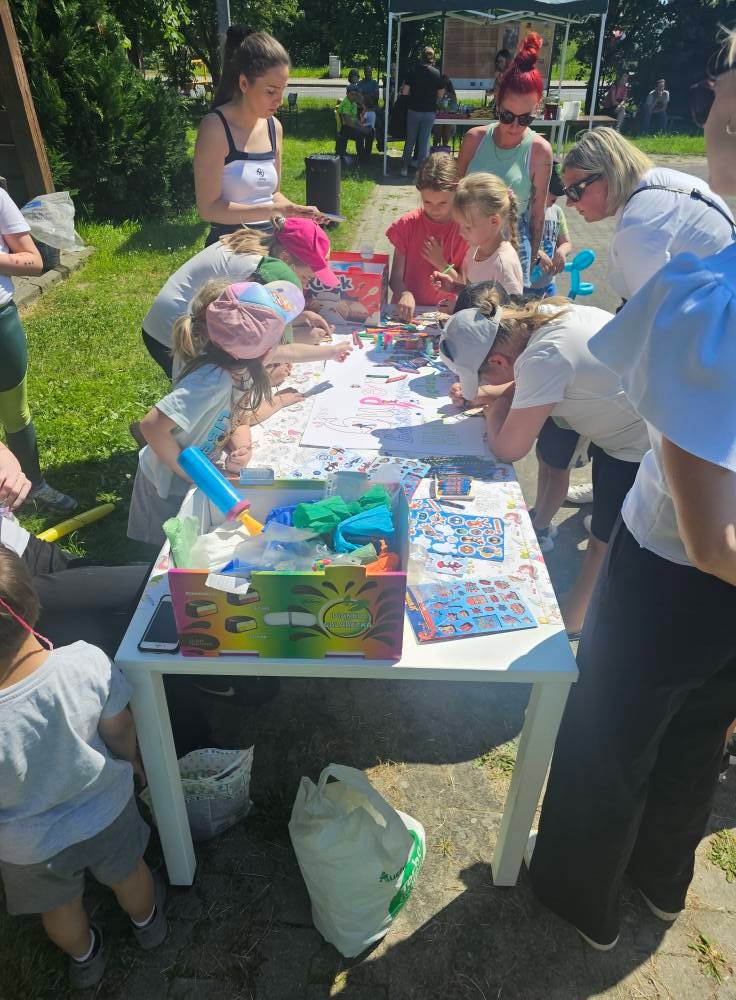 Dzieci rysują na długim stole pod opieką dorosłych podczas eventu plenerowego. Widoczne kredki, naklejki i akcesoria plastyczne. Jasne oświetlenie.