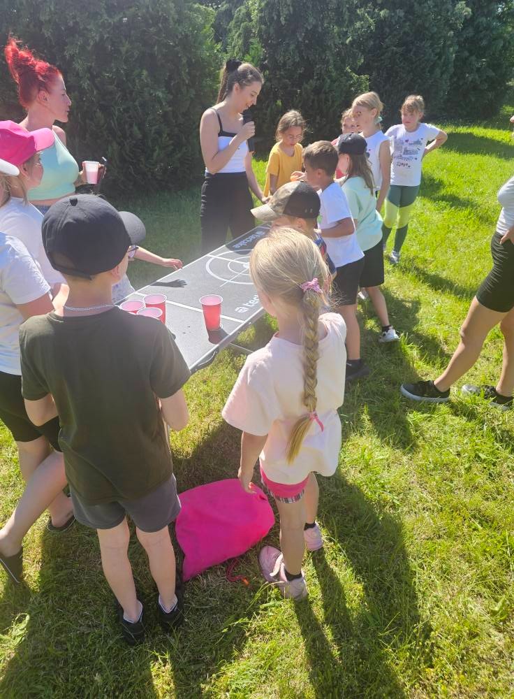 Dzieci grają w beer pong na trawie, otoczone dorosłymi i zielenią. Czerwone kubki i gra na stole z tarczą. Jasne światło słoneczne.