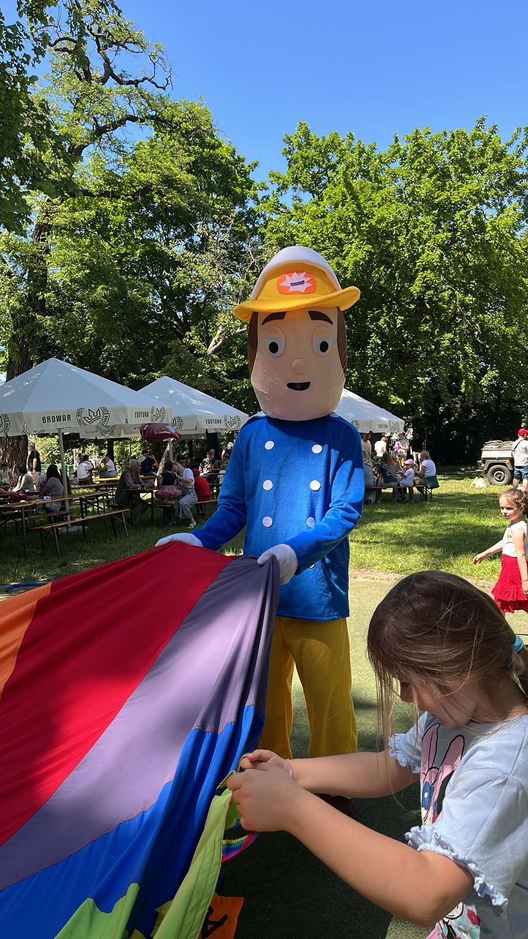 Animator w stroju strażaka Sama bawi się z dziećmi na pikniku, trzymając kolorową chustę animacyjną. W tle widać parasole i ludzi.