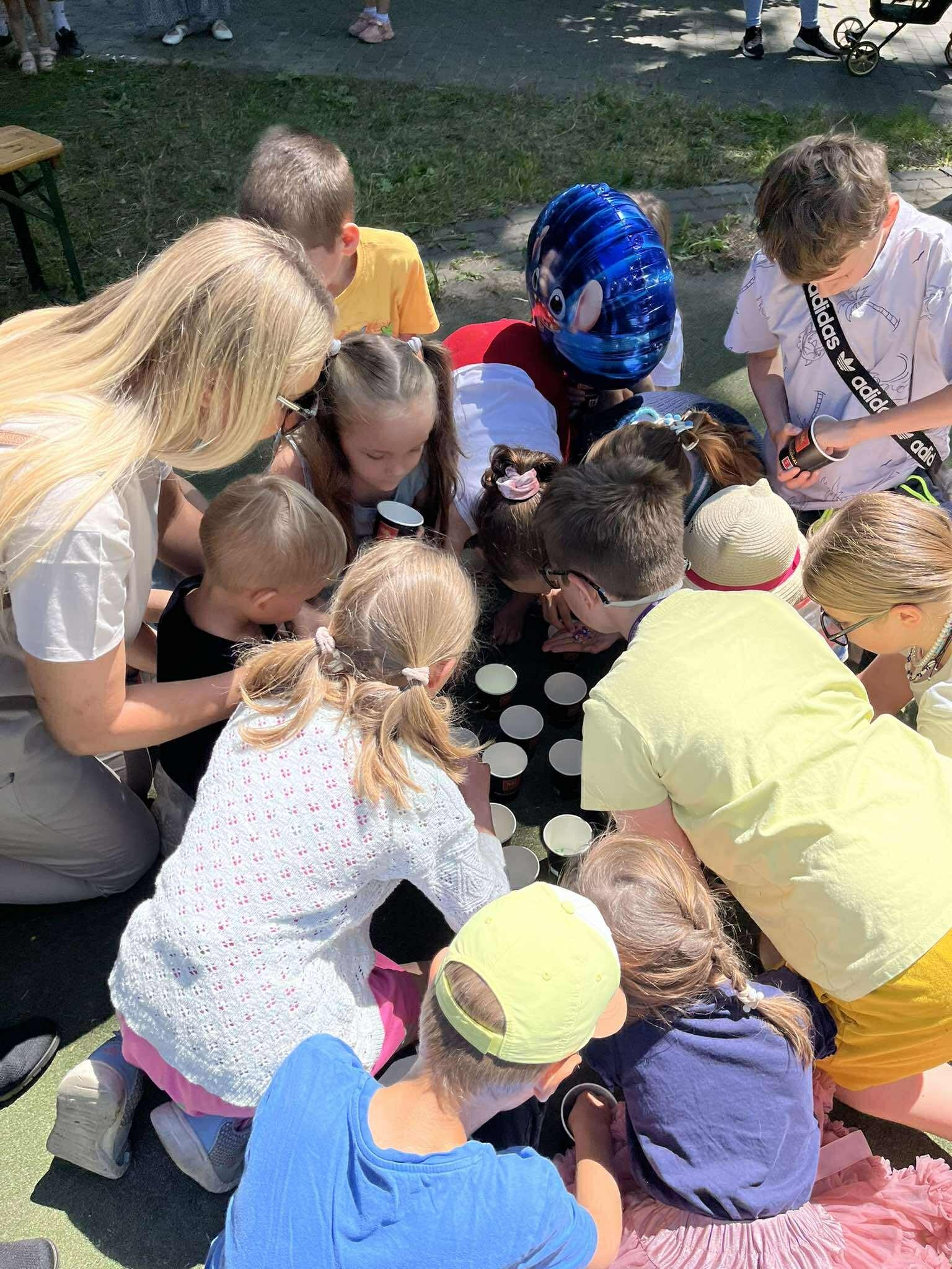 Grupa dzieci i dorosły pochyleni nad rzędem kubeczków podczas zabawy na świeżym powietrzu. Widoczny fragment zielonej powierzchni i ubrania uczestników.