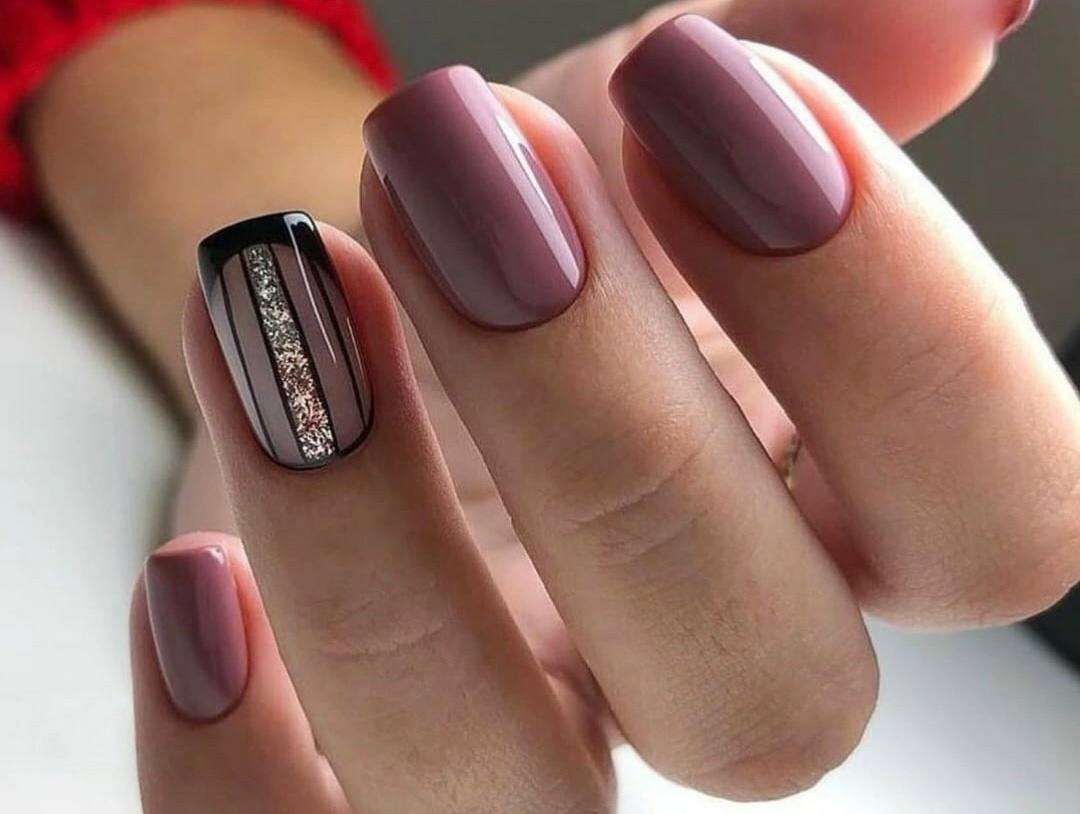 Dłoń z manicure w odcieniach mauve, jeden paznokieć z czarnym wykończeniem i srebrnym brokatowym paskiem pośrodku, pozostałe paznokcie w jednolitym kolorze.