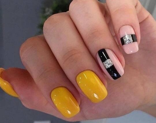 Dłoń z pomalowanymi paznokciami: dwa żółte, jeden różowy z czarnym paskiem i srebrnymi zdobieniami, jeden różowy z czarnym paskiem i srebrnymi zdobieniami.