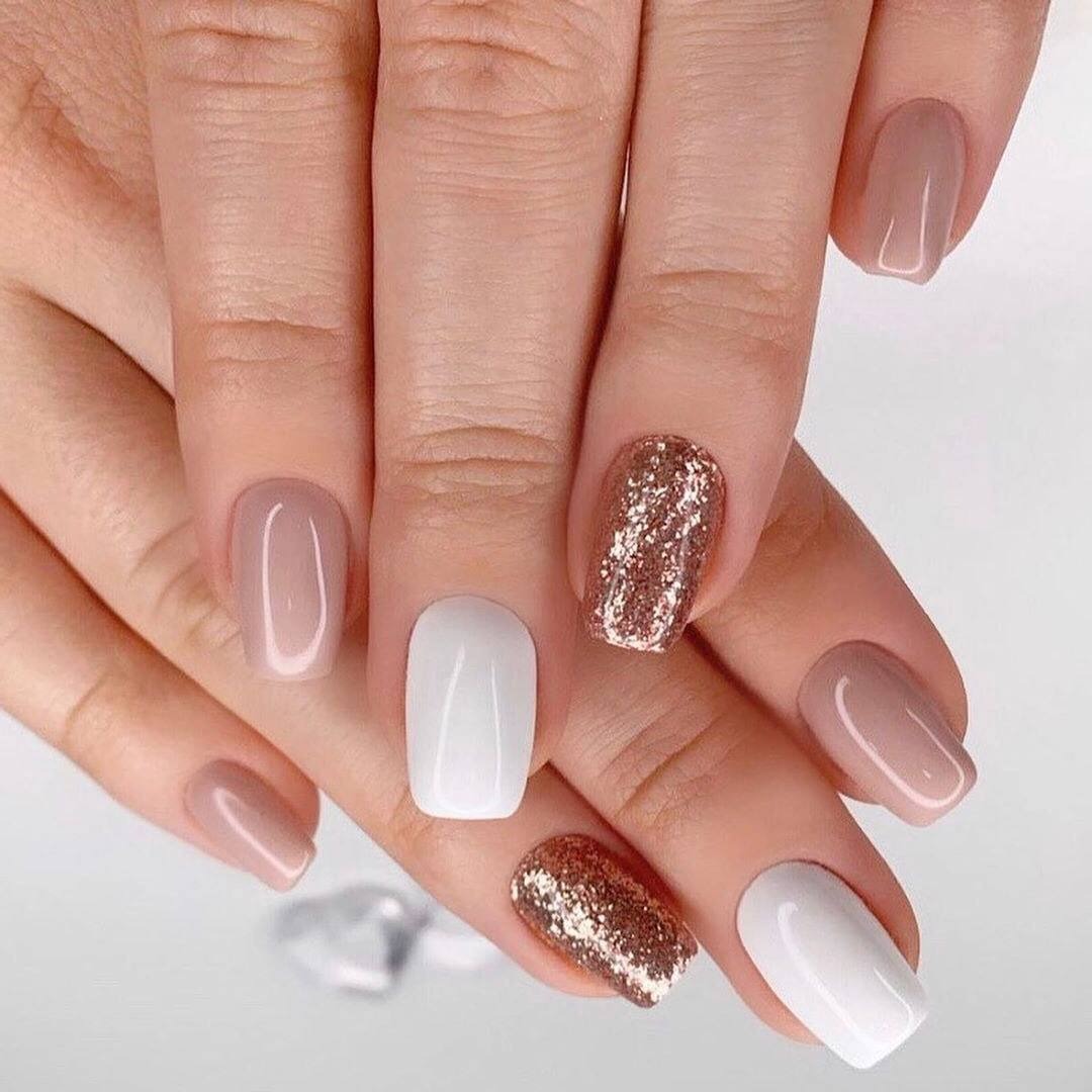 Dłonie prezentują stylizację paznokci: trzy paznokcie pomalowane na beżowo, dwa na biało, a dwa pokryte brokatem w odcieniach złota i miedzi na krótkich, kwadratowych płytkach.