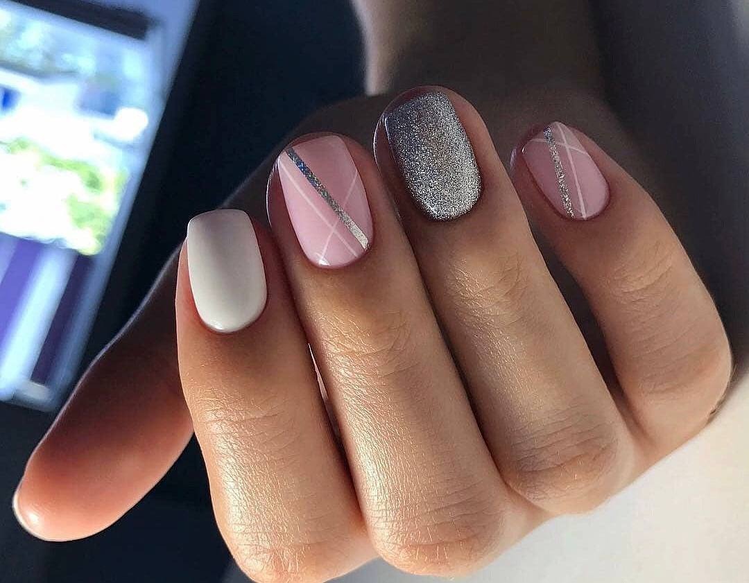 Dłoń z manicure: jeden paznokieć biały, dwa jasnoróżowe z srebrnymi paskami, jeden srebrny brokatowy.