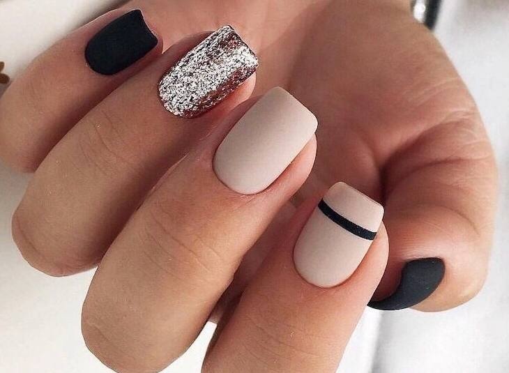 Dłoń z pomalowanymi paznokciami: jeden czarny matowy, jeden srebrny brokatowy, jeden beżowy matowy, jeden beżowy z czarnym paskiem, drugi czarny matowy.