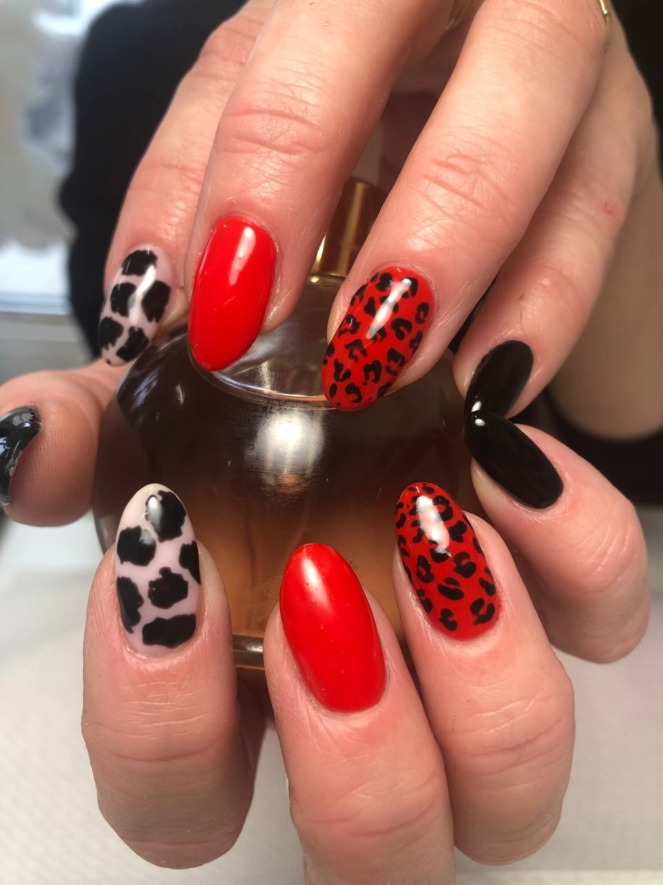 Dłonie z pomalowanymi paznokciami w różne wzory: krowie łaty, panterka i jednolity czerwony oraz czarny lakier, na tle flakonu perfum.