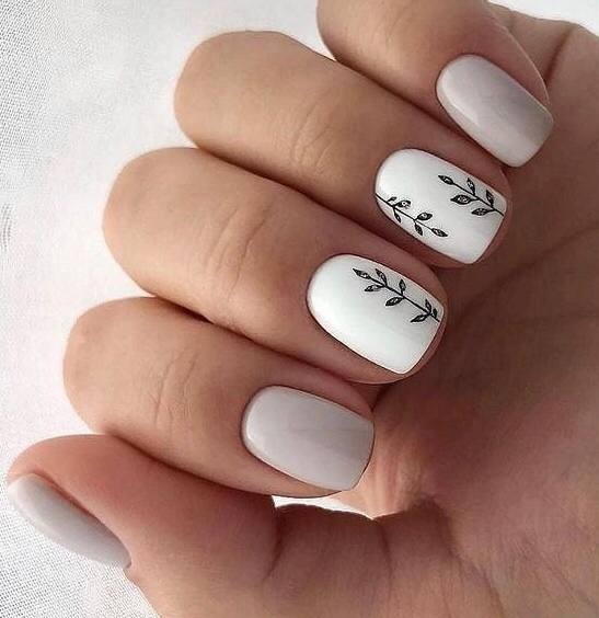 Dłoń z pomalowanymi paznokciami w odcieniach bieli i szarości, dwa paznokcie ozdobione drobnymi, czarnymi rysunkami gałązek z listkami.