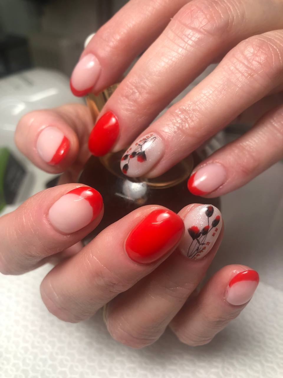 Dłonie z pomalowanymi paznokciami: klasyczny french manicure z czerwonymi końcówkami i jeden paznokieć z delikatnym, kwiatowym wzorem na jasnym tle, trzymające butelkę olejku do skórek.