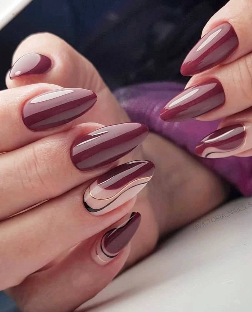 Dłonie z bordowymi paznokciami w kształcie migdała, zdobionymi złotą i czarną linią na beżowym tle, połyskujące w świetle.