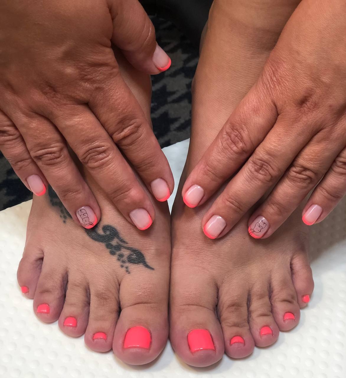 Dłonie i stopy z french manicure w neonowym koralowym odcieniu, na palcu stopy tatuaż z motywem kwiatowym.