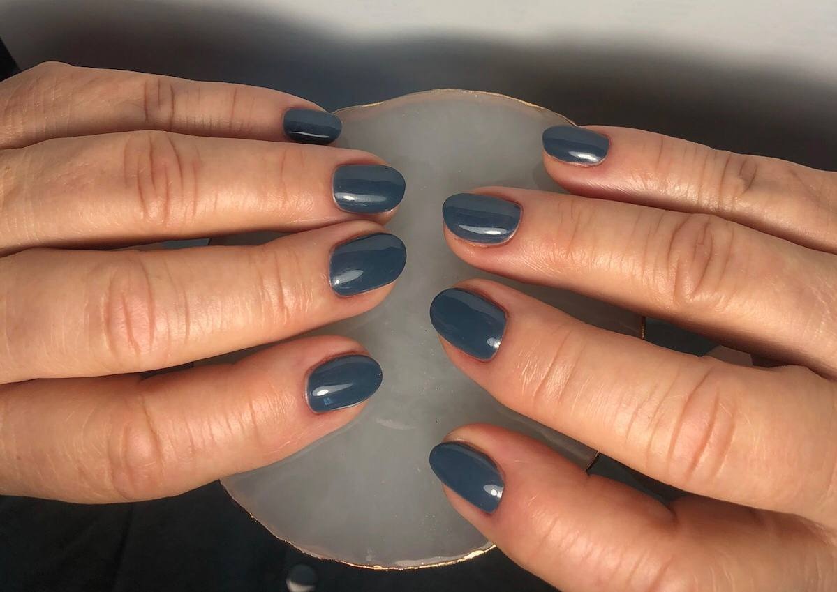 Dłonie z krótkimi, pomalowanymi na ciemny, szary kolor paznokciami, ułożone na jasnym, okrągłym podkładzie z delikatnym złotym obramowaniem.