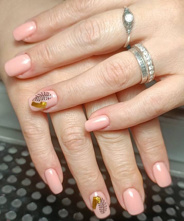 Dłonie z delikatnym, różowym manicure z roślinnym zdobieniem w kolorze złota i czerni na dwóch paznokciach; widoczne pierścionki na palcach.