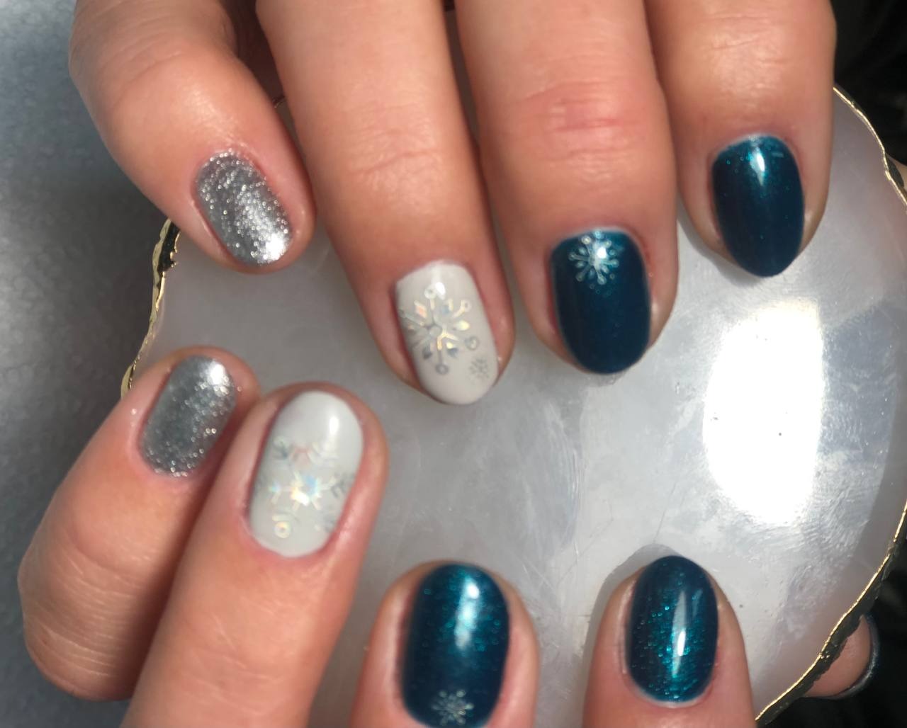 Zimowy manicure: paznokcie w kolorach srebrnym, białym i ciemnoturkusowym, zdobione motywem śnieżynek, prezentowane na tle marmurkowej podkładki.