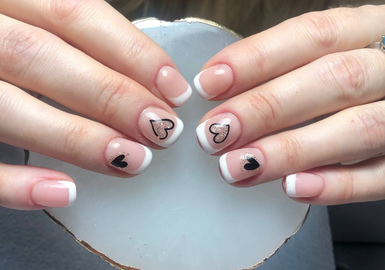 Dłonie z krótkimi paznokciami w stylu french z czarnymi i brokatowymi serduszkami na beżowym tle, ułożone na okrągłej, jasnej podkładce z delikatnym złotym obramowaniem.