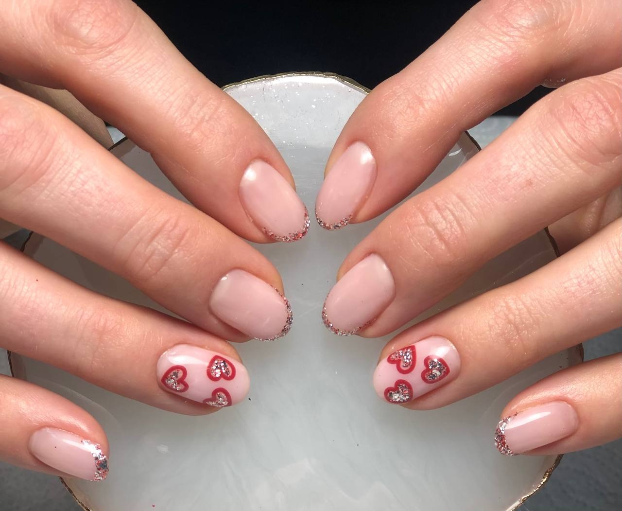 Delikatny, różowy manicure hybrydowy z brokatowymi końcówkami i wzorem czerwonych serduszek z brokatowym obrysem na dwóch paznokciach, ułożone na białym podłożu.