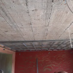 Surowy betonowy strop z widocznymi śladami szalunku, metalowa konstrukcja podwieszanego sufitu, czerwona ściana z dekoracyjnym wzorem i instalacja elektryczna w trakcie prac wykończeniowych.