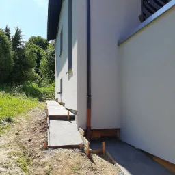 Świeżo wylany betonowy chodnik przy ścianie budynku z widocznym deskowaniem, grunt wokół nieuporządkowany.