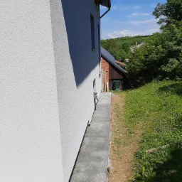 Świeżo otynkowana elewacja domu jednorodzinnego w Przemyślu, z widocznym fragmentem betonowego chodnika wokół budynku i zielonym otoczeniem.