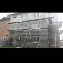 Budynek wielorodzinny w trakcie prac elewacyjnych, z widocznymi rusztowaniami, nałożonym szarym tynkiem i oknami. Obok widoczna ściana z czerwonej cegły innego budynku.