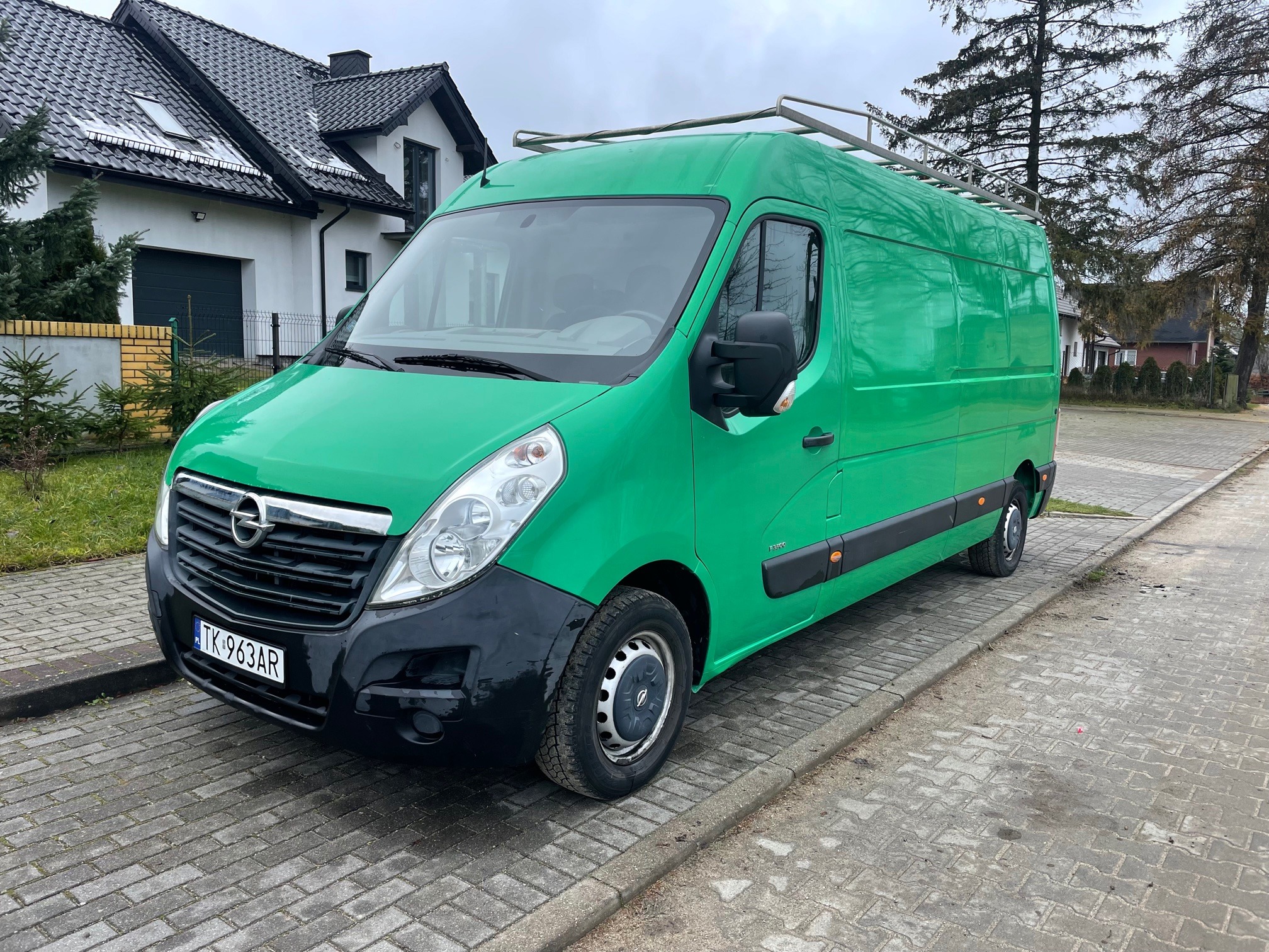 Zielony Opel Movano z relingami dachowymi zaparkowany na chodniku z kostki brukowej przed domem jednorodzinnym w pochmurny dzień.