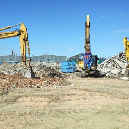 Przedsiębiorstwo CAMA Sp. z o.o. - Trzy żółte koparki Komatsu na placu budowy z gruzem i pracownikiem w pomarańczowej kamizelce, widok na budynek przemysłowy w tle pod błękitnym niebem.