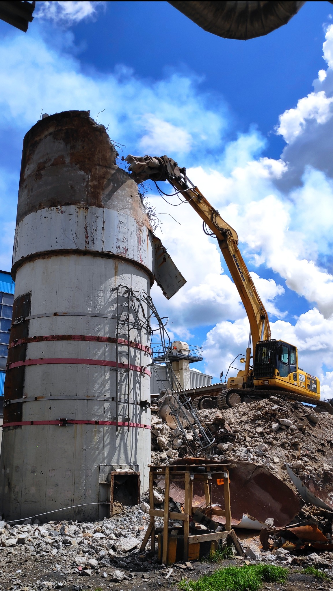 Żółta koparka Komatsu PC 290 LC z hydraulicznym młotem rozbija górną część betonowego silosu na tle błękitnego nieba z chmurami, w pobliżu gruz i metalowe elementy konstrukcyjne.