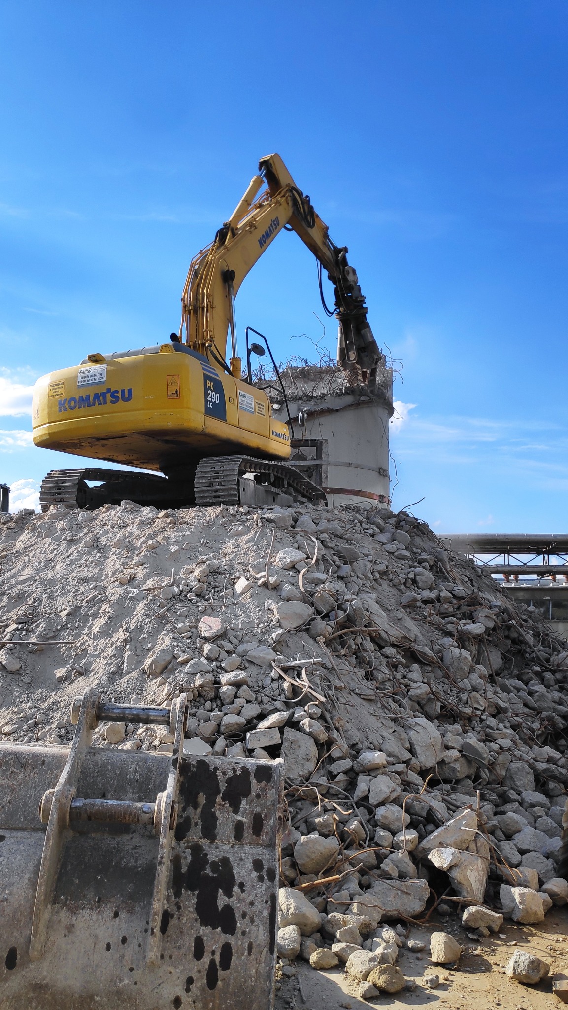 Żółta koparka Komatsu PC 290 LC na szczycie sterty gruzu, krusząca betonową konstrukcję zbrojoną stalą na tle błękitnego nieba.