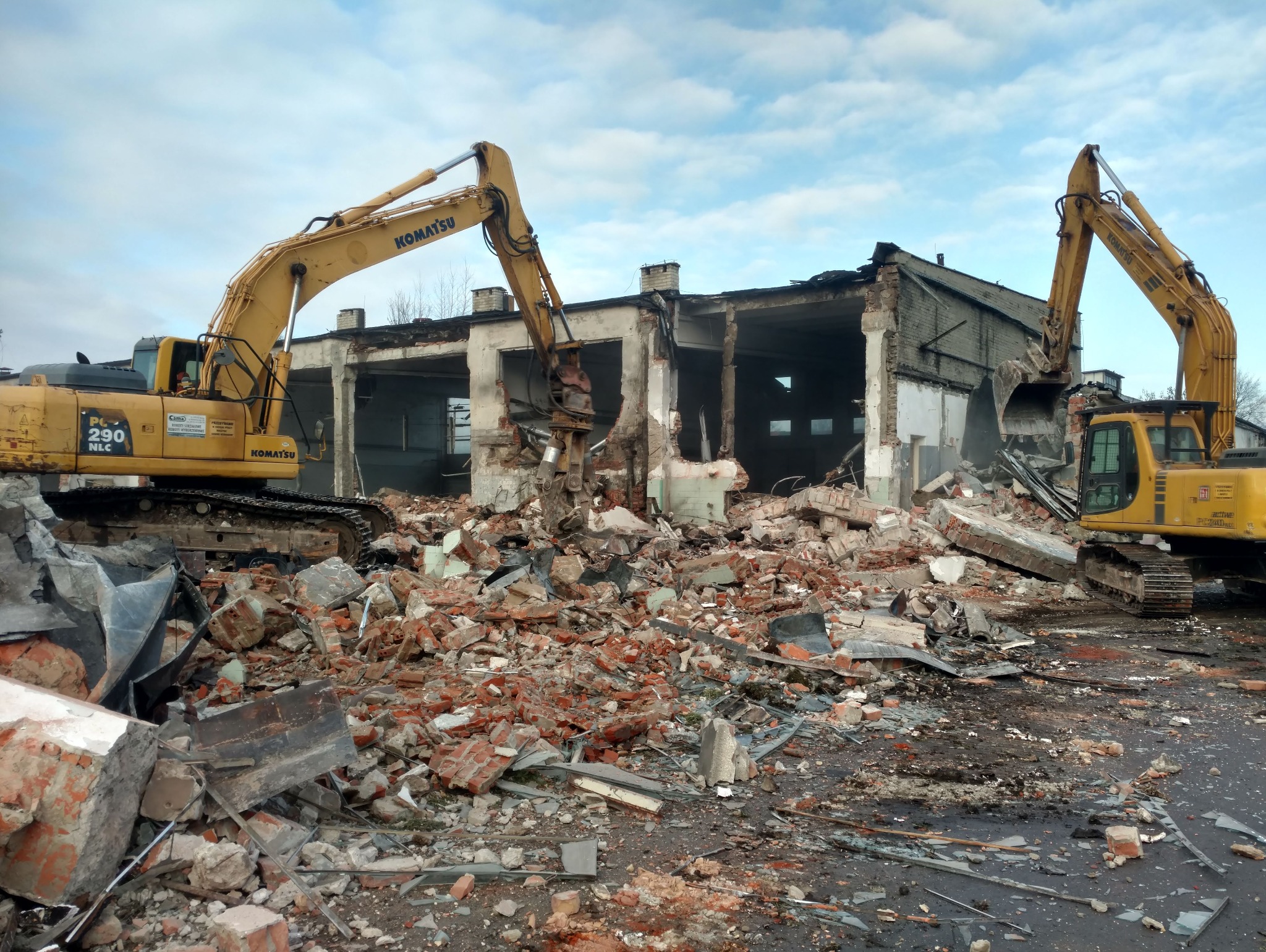 Dwa żółte koparki Komatsu pracują przy rozbiórce budynku z cegły i betonu, gruzowisko na pierwszym planie, pochmurne niebo w tle.