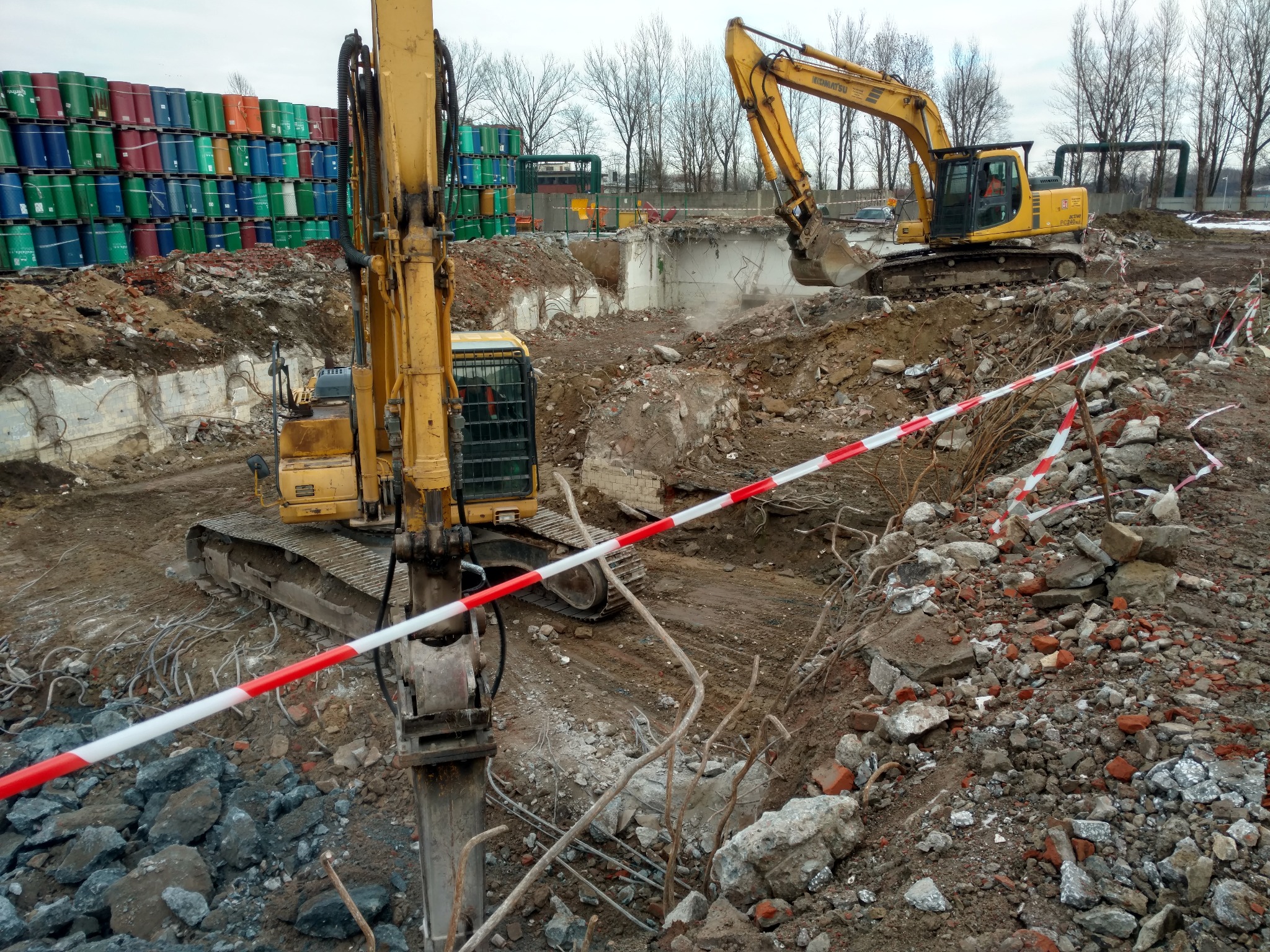Dwie żółte koparki Komatsu na placu budowy, jedna z młotem hydraulicznym, druga z łyżką, gruz i pozostałości po wyburzeniach, w tle wysokie składowisko kolorowych beczek.