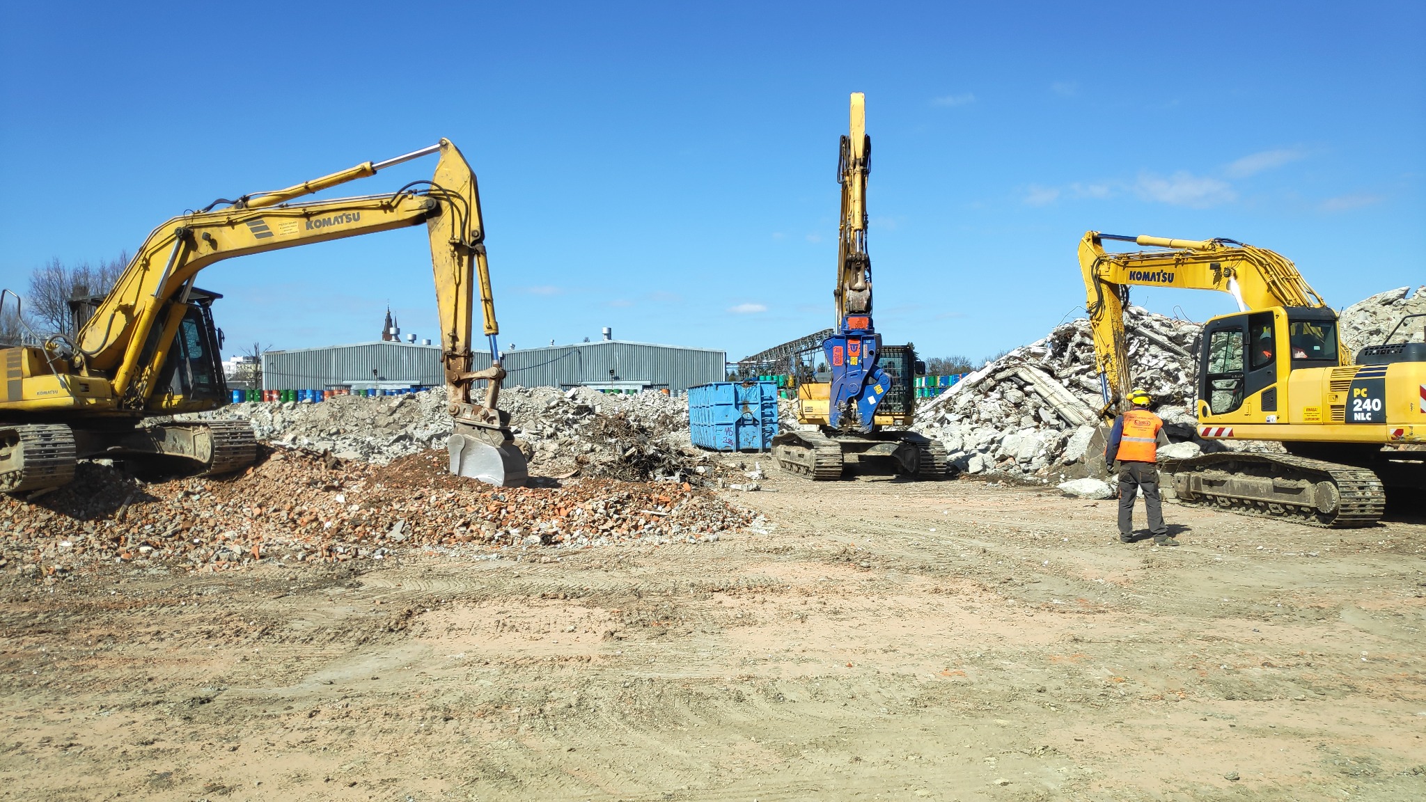 Trzy żółte koparki Komatsu na placu budowy z gruzem i pracownikiem w pomarańczowej kamizelce, widok na budynek przemysłowy w tle pod błękitnym niebem.