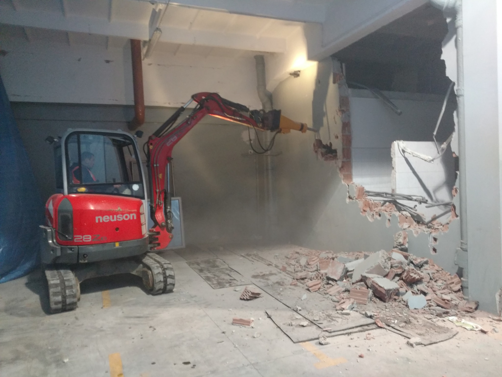 Czerwona minikoparka Neuson 28Z z hydraulicznym młotem pneumatycznym rozbija ścianę wewnątrz budynku, gruz leży na podłodze, widoczny operator w kabinie.