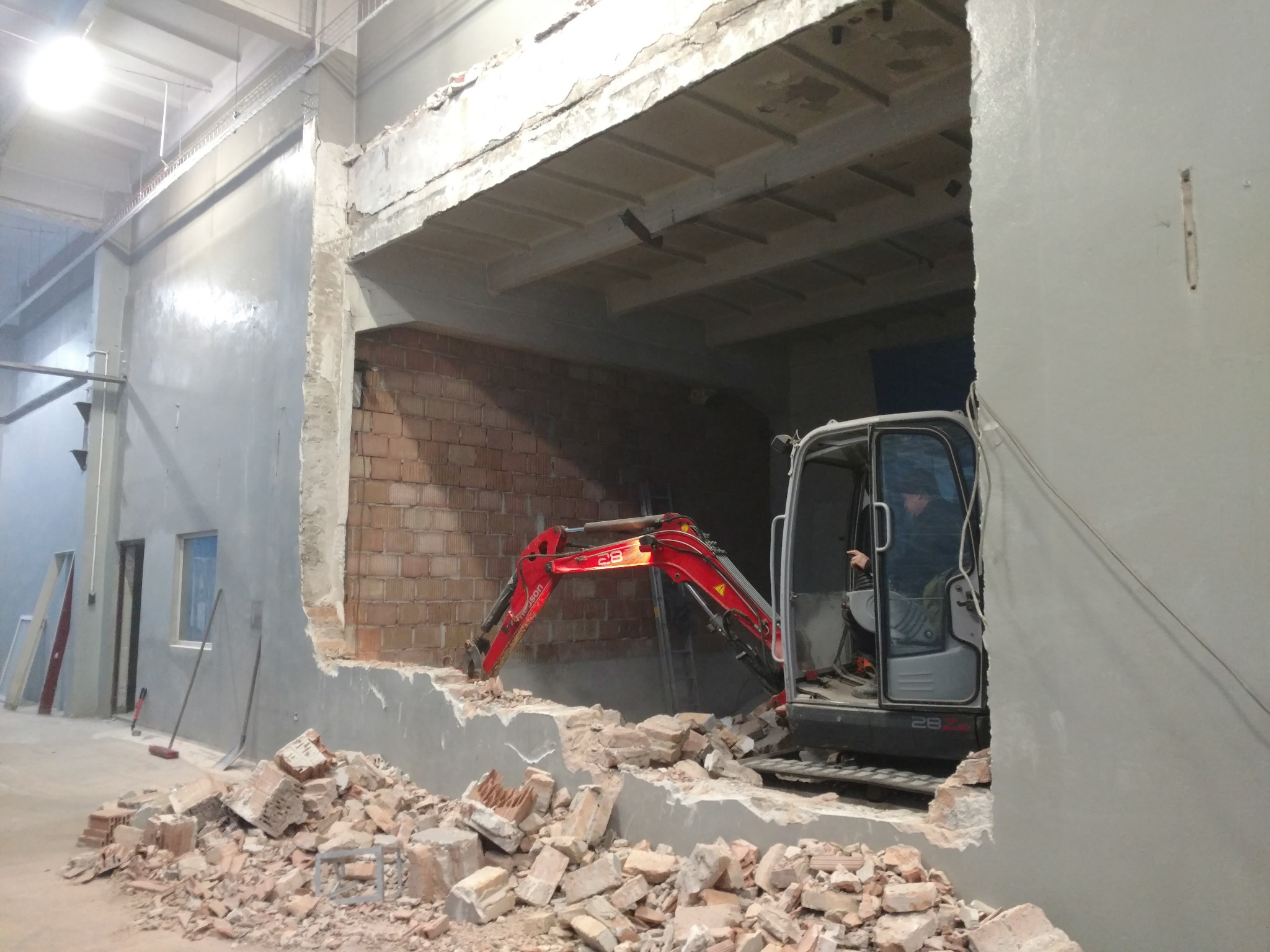 Czerwony minikoparka hydrauliczna usuwa fragment muru z cegły wewnątrz budynku przemysłowego, widoczne gruzowisko i operator w kabinie.